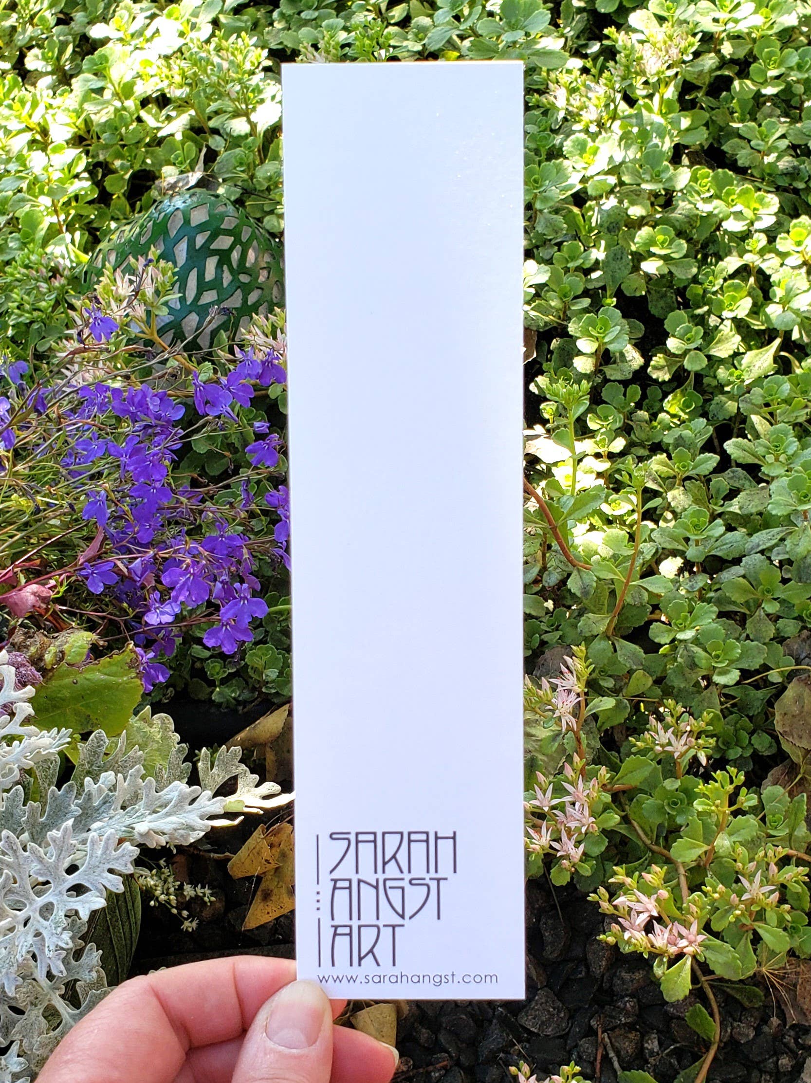 A hand holds a blank white bookmark labeled Bookmark: Mountain Bluet by Sarah Angst Art, surrounded by lush greenery and Mountain Bluet flowers. Proudly printed in the USA, this bookmark captures natures tranquility.