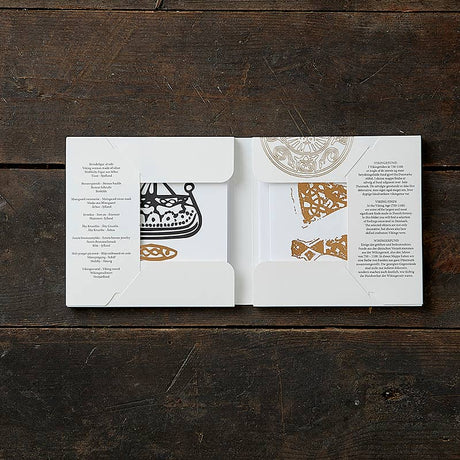 Open the Card Pack: Viking Finds Square Card Folder by Koustrup & Co - Denmark on a wooden surface to reveal intricate black and white Viking motifs with gold accents inside.