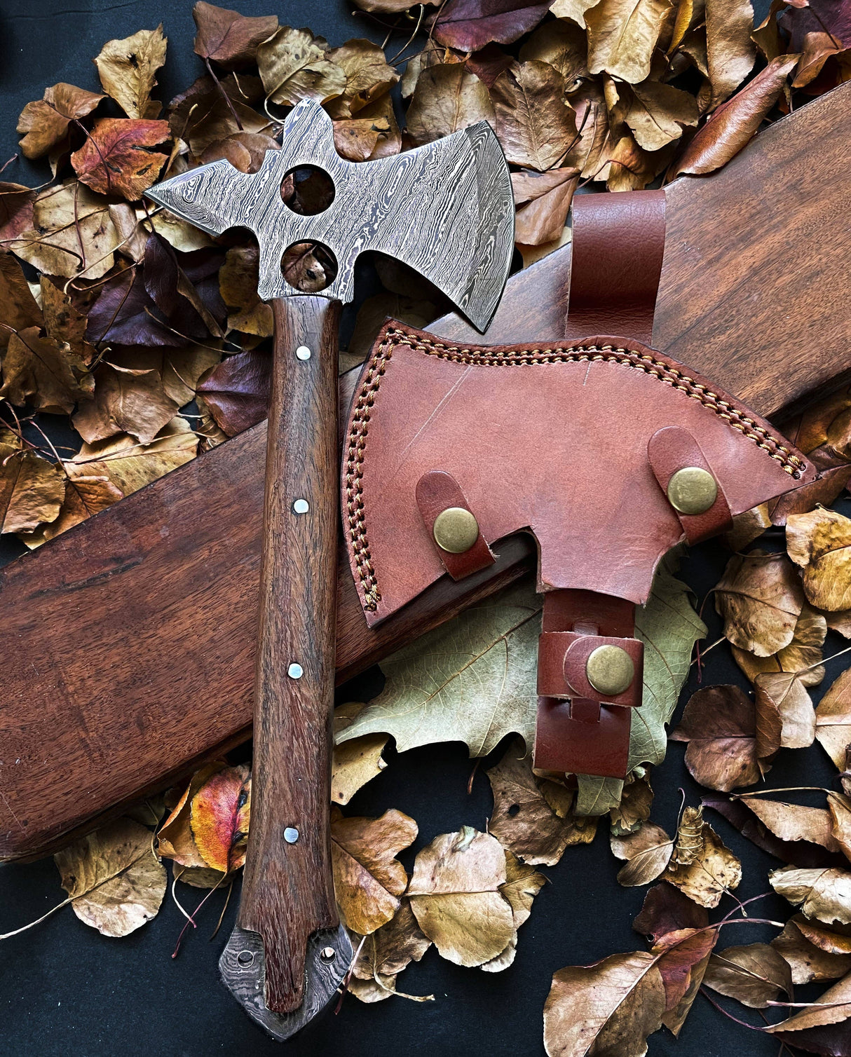 A wooden-handled Damascus tomahawk axe with a holed metal blade on fallen leaves next to a brown leather sheath featuring brass buttons and stitched edges.