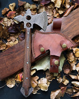 A wooden-handled Damascus tomahawk axe with a holed metal blade on fallen leaves next to a brown leather sheath featuring brass buttons and stitched edges.