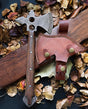 A wooden-handled Damascus tomahawk axe with a holed metal blade on fallen leaves next to a brown leather sheath featuring brass buttons and stitched edges.