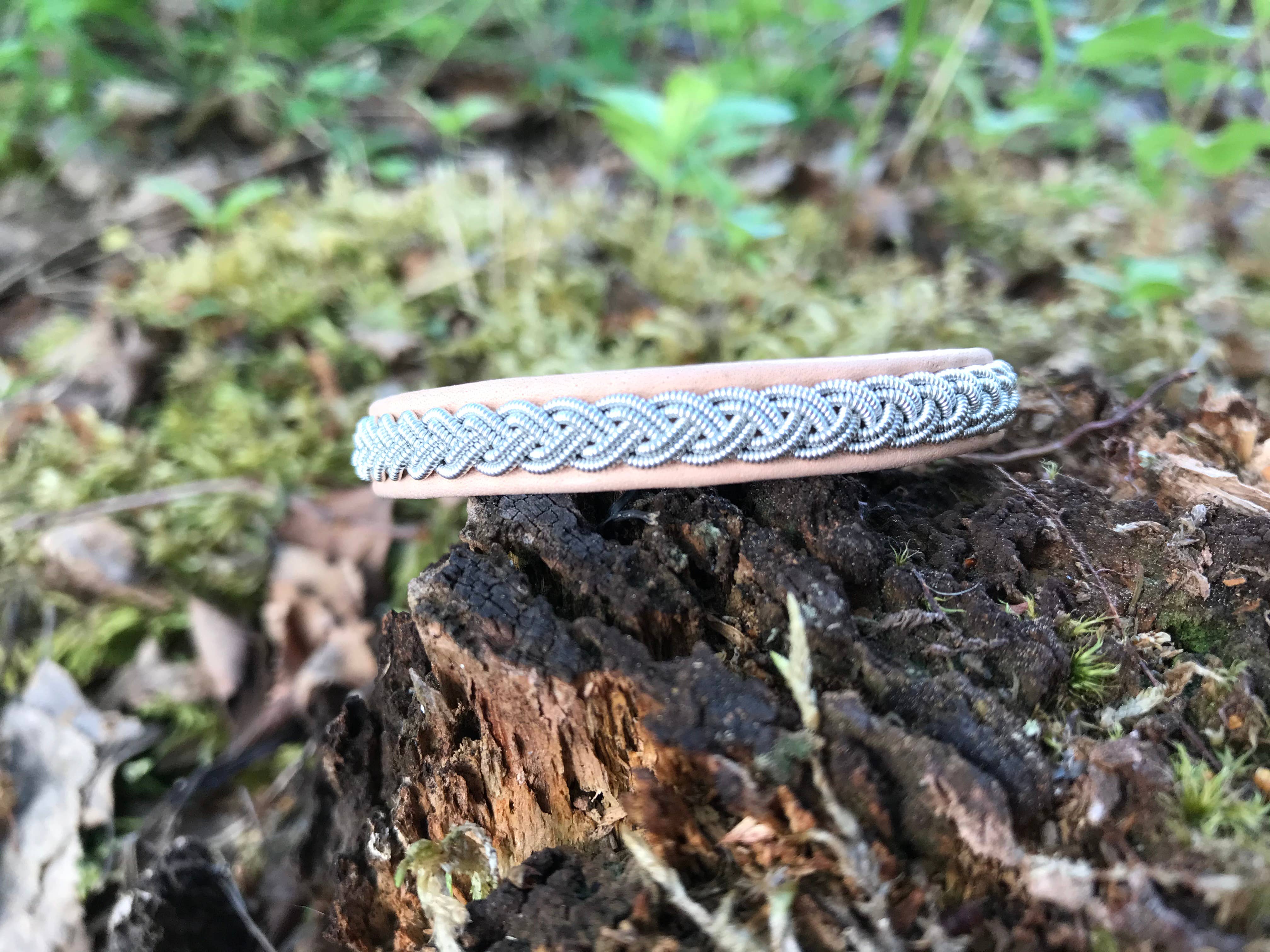 The Bracelet: Lapland Sami Tin Bracelet Single Braid #1: Blue Gray rests on a mossy tree stump outdoors, its blue-gray leather band braided with tin, surrounded by lush greenery and leaves.