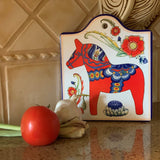 A tomato, garlic bulb, and green onion sit on a counter before the Red Dala Horse Cheeseboard by European Heritage Gifts, a ceramic wall decor piece with Swedish motifs and a hangable cork back. Tan tiles complete the cozy kitchen scene.