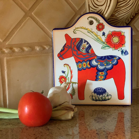 A tomato, garlic bulb, and green onion sit on a counter before the Red Dala Horse Cheeseboard by European Heritage Gifts, a ceramic wall decor piece with Swedish motifs and a hangable cork back. Tan tiles complete the cozy kitchen scene.