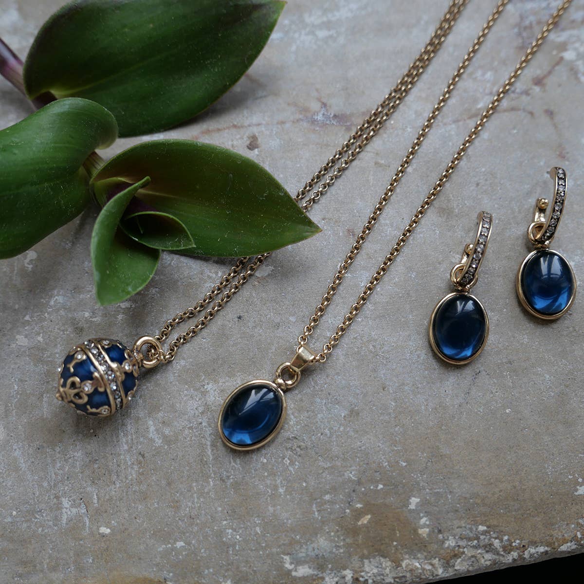 Part of the Sophistication Collection, this set features two A&C Oslo gold-plated necklaces with crystal pendants and earrings, all adorned with blue gemstones. Theyre elegantly displayed on a textured surface beside green leaves.