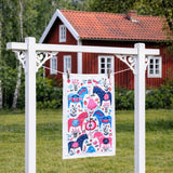 A Tea Towel: Swedish Dala Horse Linen - Cotton Kitchen Towel by Lotta Spjute hangs on a white drying rack outdoors, with a red wooden house and green trees in the background.