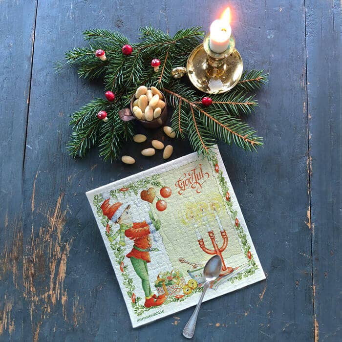 A lit candle, fir branch with berries and nuts, a festive napkin with a spoon, and the Dish Cloth: Straw Goat with Red Ribbons / Christmas are arranged on a rustic blue wooden table.