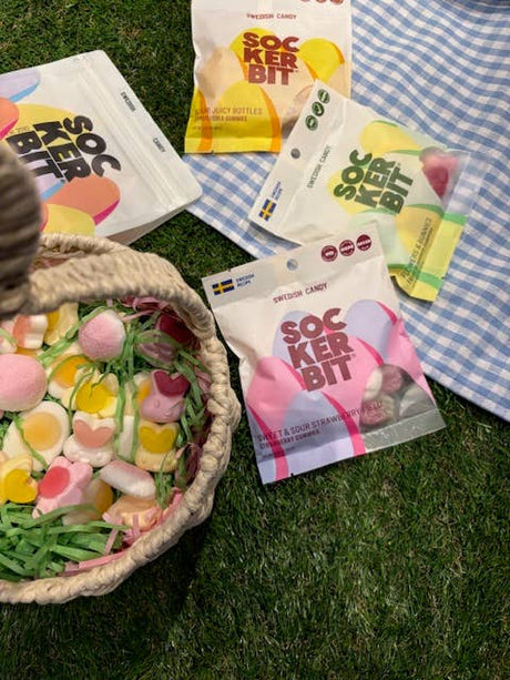 A wicker basket with colorful candy, including Sockerbit Sour Juicy Bottles Fruit Swedish Candy (2.8oz bags), sits on grass beside four open Sockerbit bags, with a blue and white checkered cloth in the corner.
