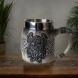 The Coffee Mug: Viking Odin's Horn Drinking Mug, featuring intricate silver patterns and a horn-shaped handle, sits on a wooden surface—an ideal Viking gift. Lush plants in the background complete this striking display.