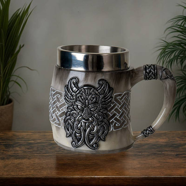 The Coffee Mug: Viking Odin's Horn Drinking Mug, featuring intricate silver patterns and a horn-shaped handle, sits on a wooden surface—an ideal Viking gift. Lush plants in the background complete this striking display.