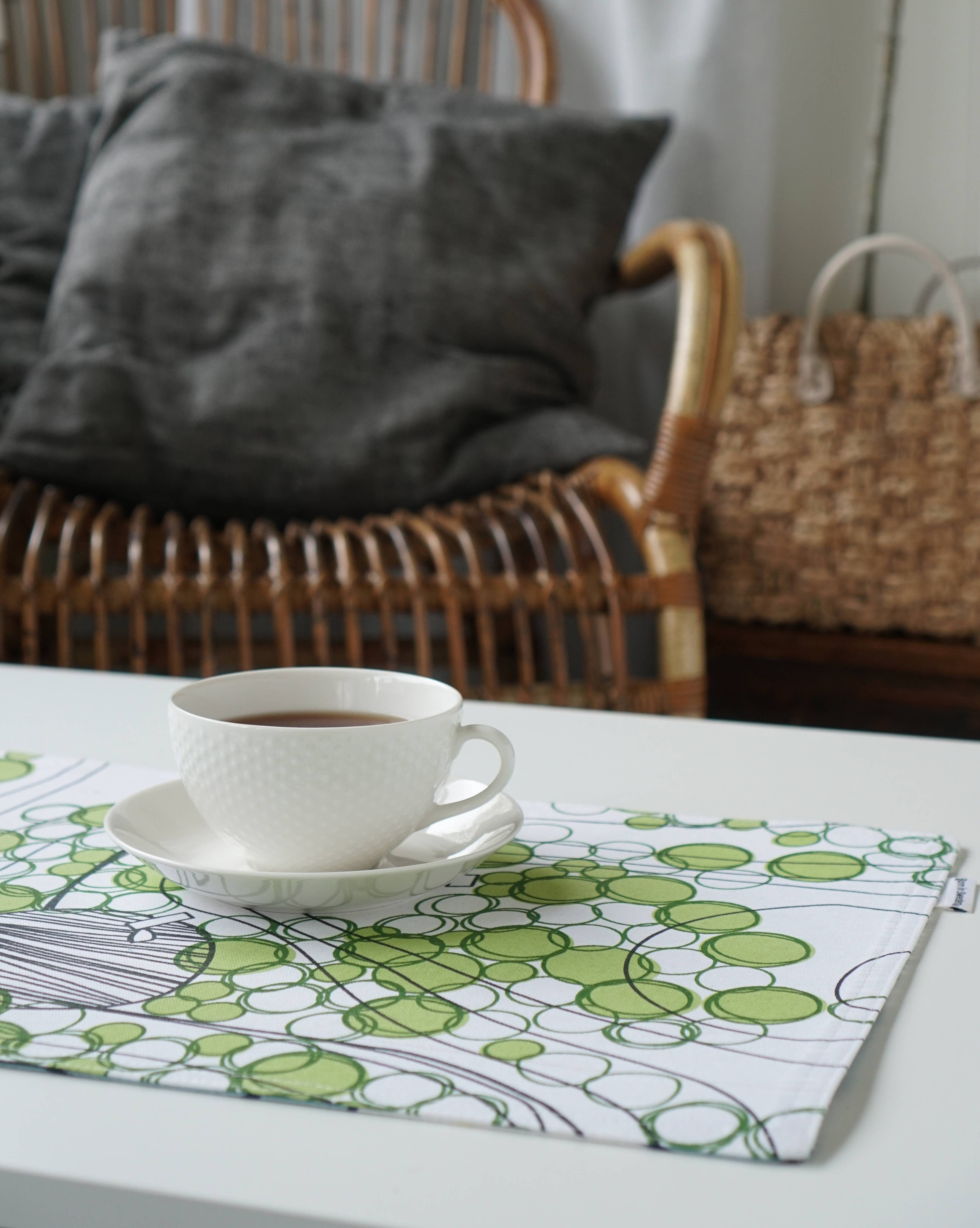 A white cup of tea on a saucer rests on the Born in Sweden double-sided cotton placemat, measuring 47 cm by 34 cm and featuring a green and white pattern. In the background, a wicker chair with gray cushions and a woven basket adds to the serene setting.