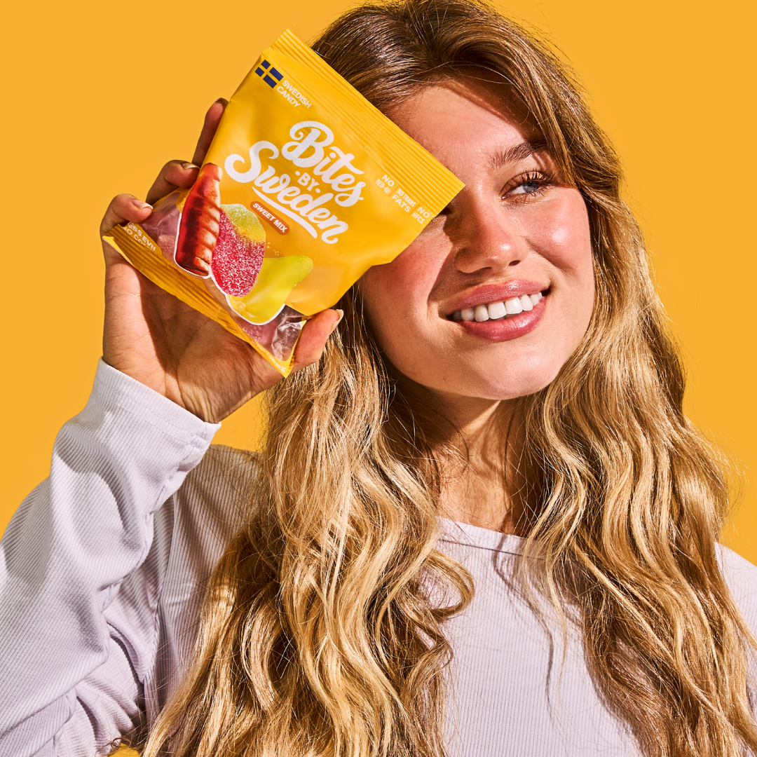A woman with long blonde hair holds a yellow Bites of Sweden candy bag next to her face, standing against a solid yellow background.
