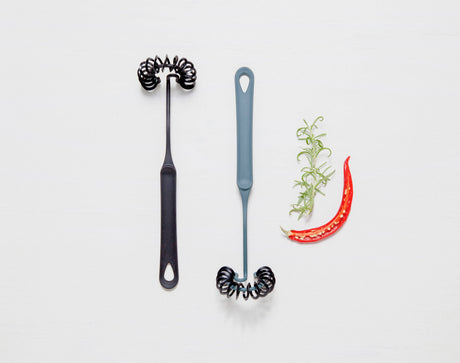 Two black Gourmaid Whisks with spiral ends, made from BPA-free, heat-resistant silicone, are placed beside sliced red chili and fresh rosemary on a white background.