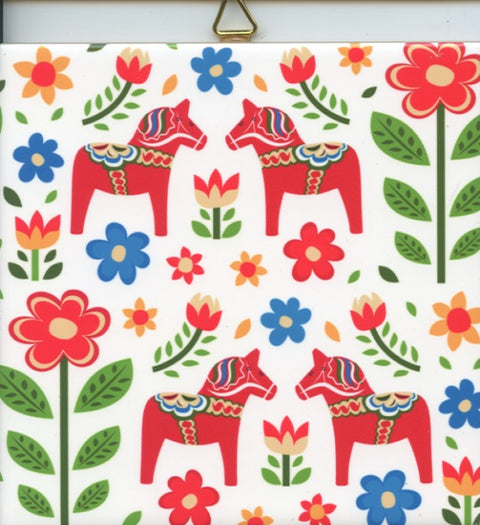 This 6" tile, named "Tile: Dala Horses & Flowers," showcases a colorful Scandinavian pattern with red Dala Horses and vibrant flowers, complemented by a white ceramic surface and cork backing. It also features a hanger for easy display.