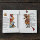 An open folder shows two cards from the "Jul A taste of Christmas" Koustrupco pack (8 cards & envelopes, 8 different images) featuring Scandinavian food illustrations and printed text, on a wooden surface.