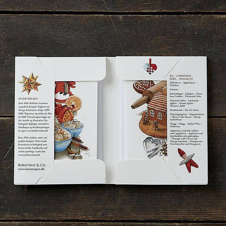 An open folder shows two cards from the "Jul A taste of Christmas" Koustrupco pack (8 cards & envelopes, 8 different images) featuring Scandinavian food illustrations and printed text, on a wooden surface.