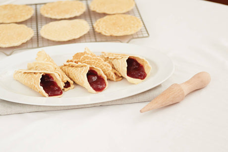 Rolled pizzelle cookies filled with red jam rest on a plate beside a Beechwood Krumkake Cone Roller (1.5" x 1.5" x 8.25"), while flat pizzelle cookies cool on a rack in the background.