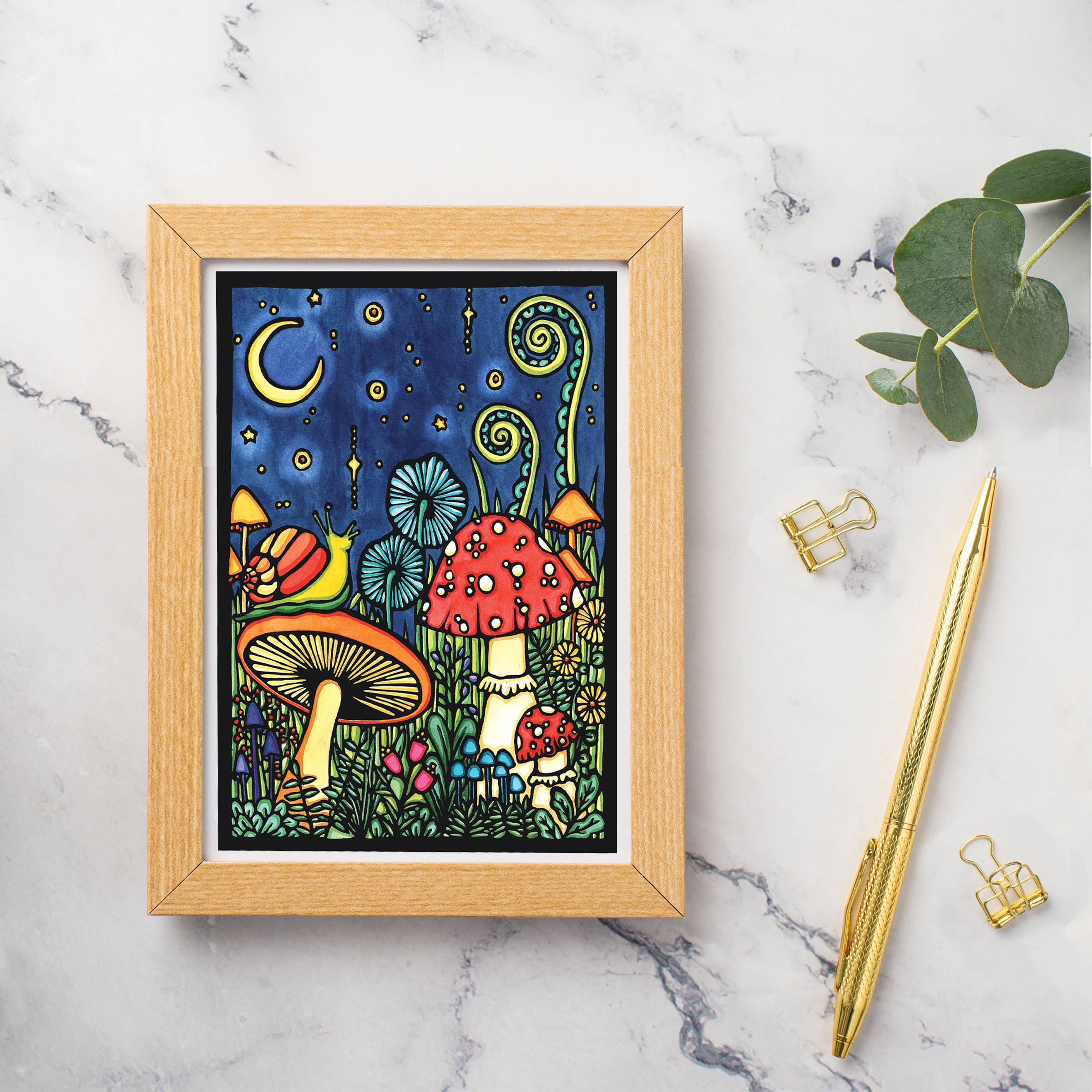 Sarah Angst Arts framed artwork, inspired by the Mushroom Greeting Card, beautifully showcases colorful mushrooms, stars, and a crescent moon on a marble surface. Complemented by a gold pen, two gold binder clips, and green leaves, it creates an enchanting visual experience.