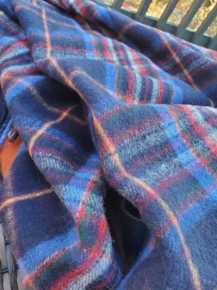 A close-up of the Esprit du Quebec Deluxe Highland Blanket, featuring blue, red, and green Quebec tartan from the Highland Collection, draped over a surface with some lint on its soft plaid wool fabric.