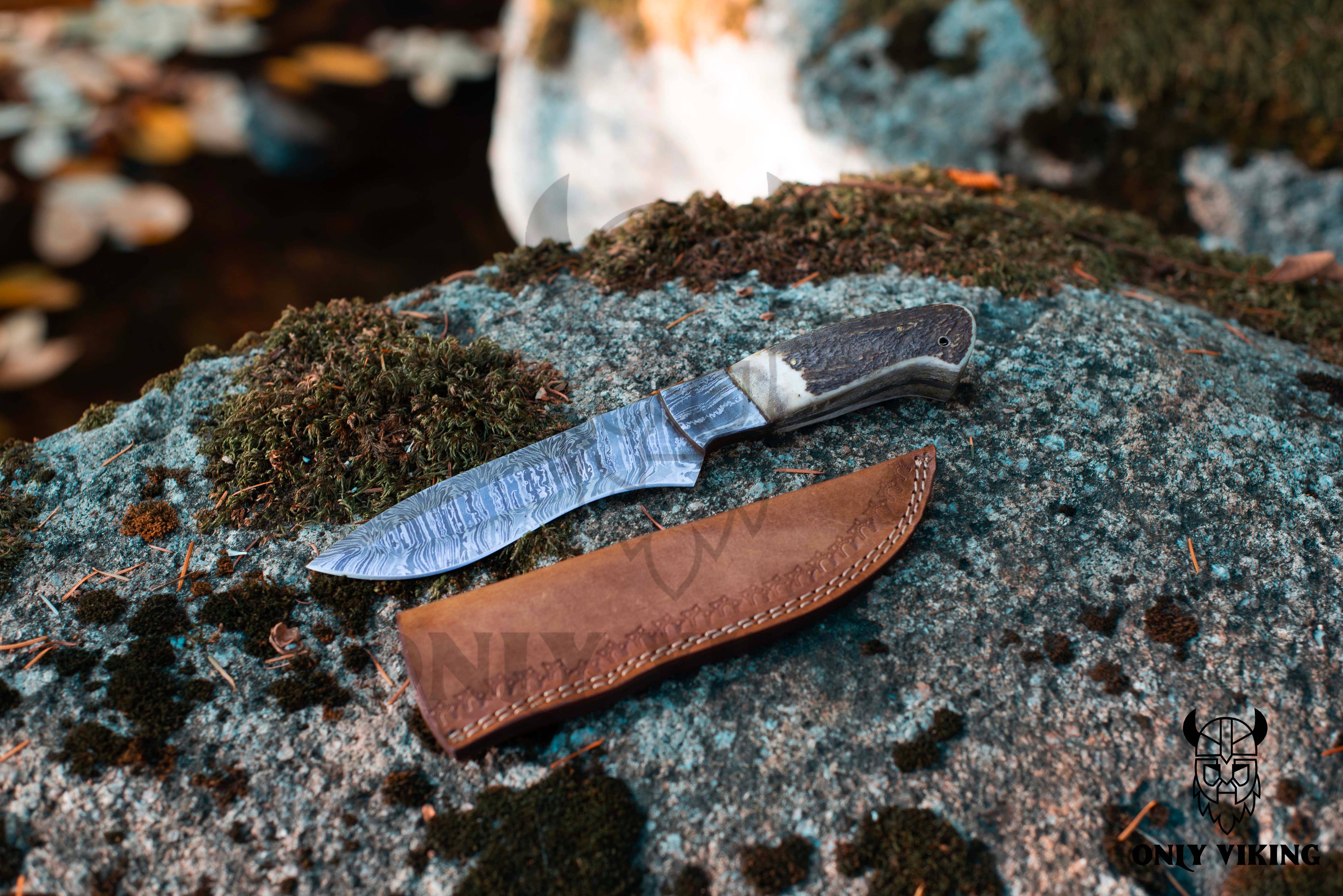 A Knife: Damascus Hunting Knife With Leather Sheath, featuring a textured blade and dark handle, rests on moss-covered rock alongside its brown leather sheath, ideal for outdoor activities.