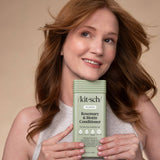 Smiling woman with long brown hair holds a green bottle of Hair: Volumizing Rosemary & Biotin Conditioner 12 fl oz. in front of her chest against a beige background, highlighting this thickening and volumizing conditioner.