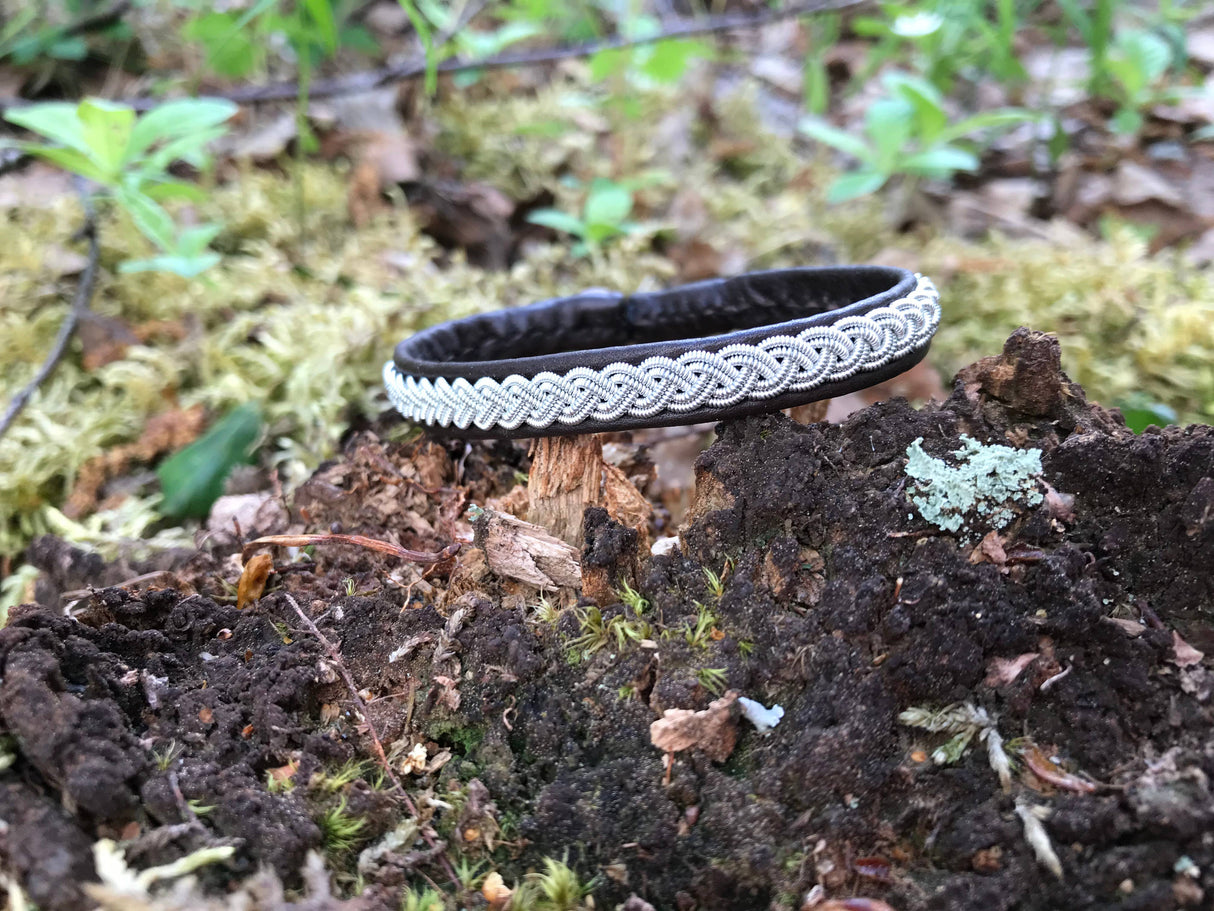 The Bracelet: Traditional Sami Single Braid Bracelet - Black #1, handcrafted in Lapland, Sweden, lies on a mossy tree stump among soil and green plants in a tranquil forest setting.
