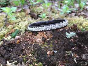 The Bracelet: Traditional Sami Single Braid Bracelet - Black #1, handcrafted in Lapland, Sweden, lies on a mossy tree stump among soil and green plants in a tranquil forest setting.