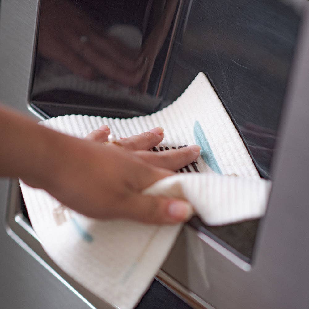 A hand wipes a stainless steel surface with a white cloth, demonstrating the effectiveness of the "Life is Better at the Lake" Swedish Dish Cloth, made from sustainable raw materials.