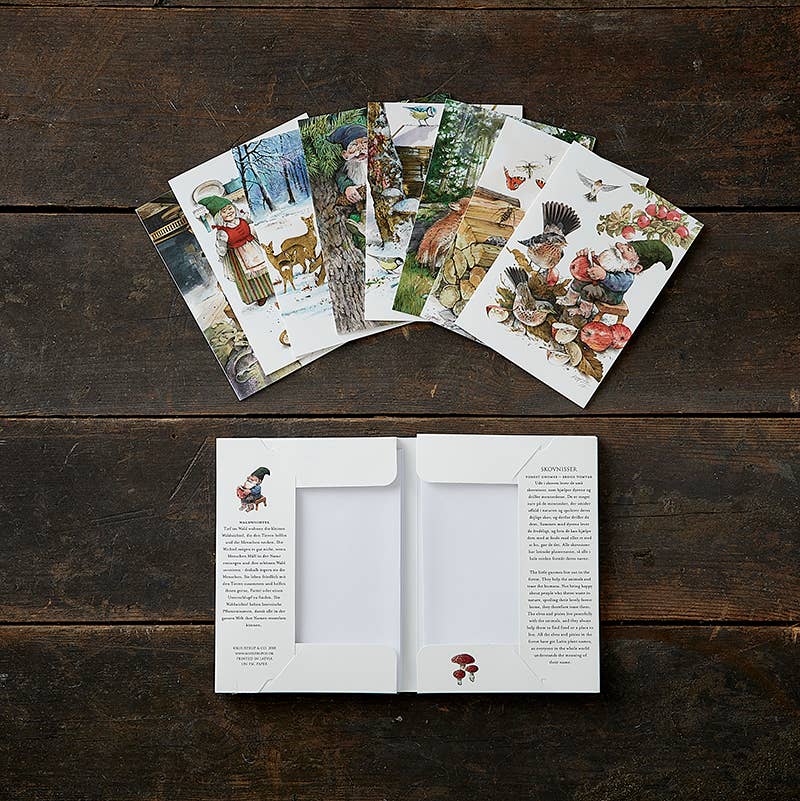 A set of illustrated "Card Pack: Forrest Gnomes - Skovnisser" postcards, featuring eight different cards with nature themes, is spread out on a wooden surface alongside an open folder for easy viewing.