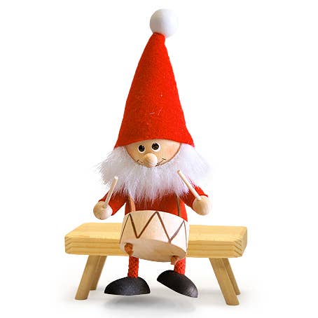 The Santa Gnome Playing Drum figurine features a handcrafted Scandinavian design with a red hat, white beard, and sits on a wooden bench while playing a drum.