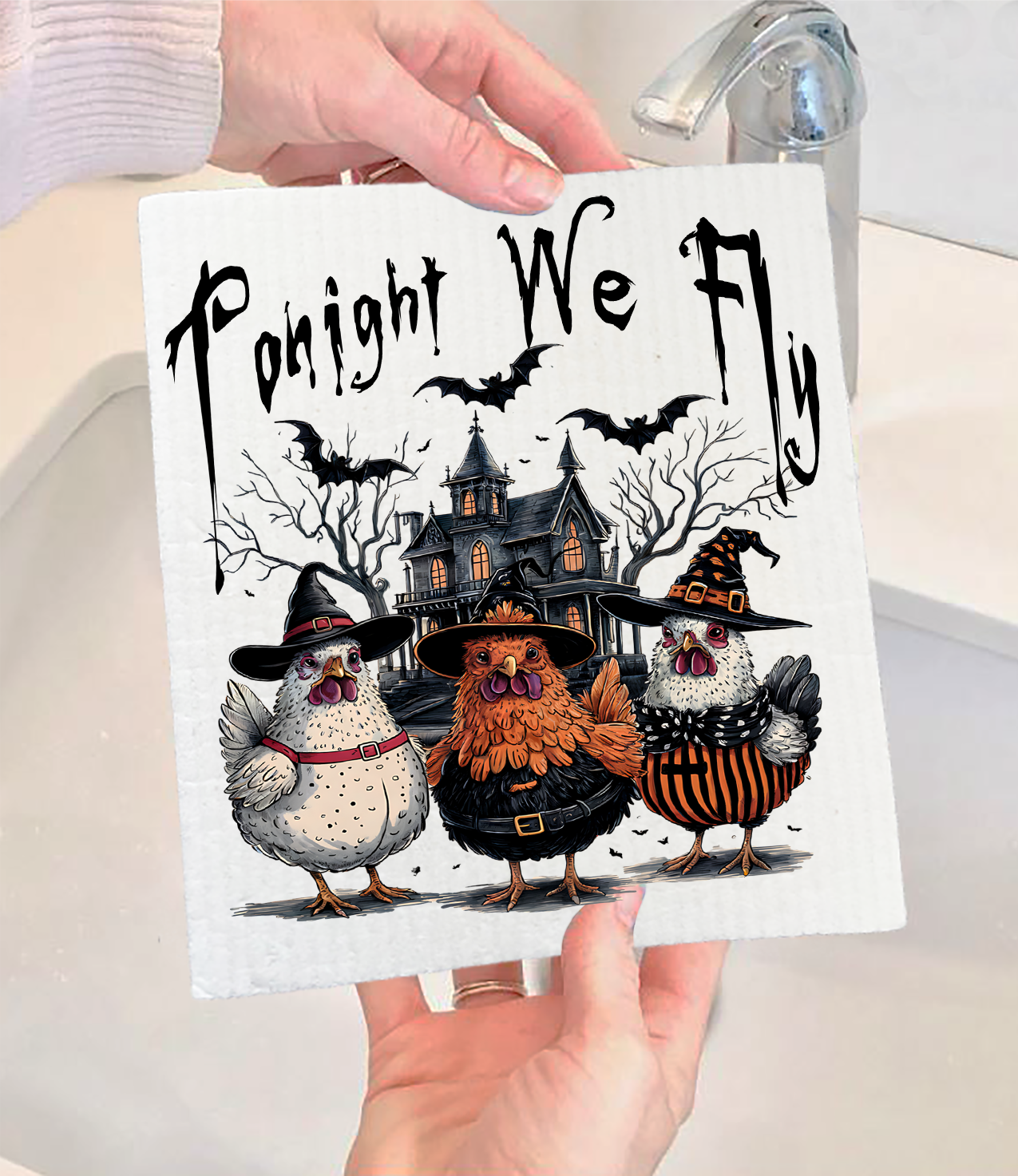 A person holds the "Dish Cloth: Halloween Chickens Tonight We Fly Swedish Dishcloth," which features three chickens in witch costumes, a haunted house, bats, and the phrase “Tonight We Fly.”.