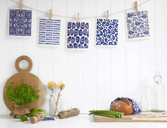 In a cozy kitchen scene exuding Scandinavian style, patterned cards hang on a line above a wooden cutting board adorned with fresh herbs. A wrapped loaf of bread rests amid asparagus, glass bottles, and spools of twine, all perfectly complemented by the eco-friendly "Robins Blue" biodegradable dish cloths.