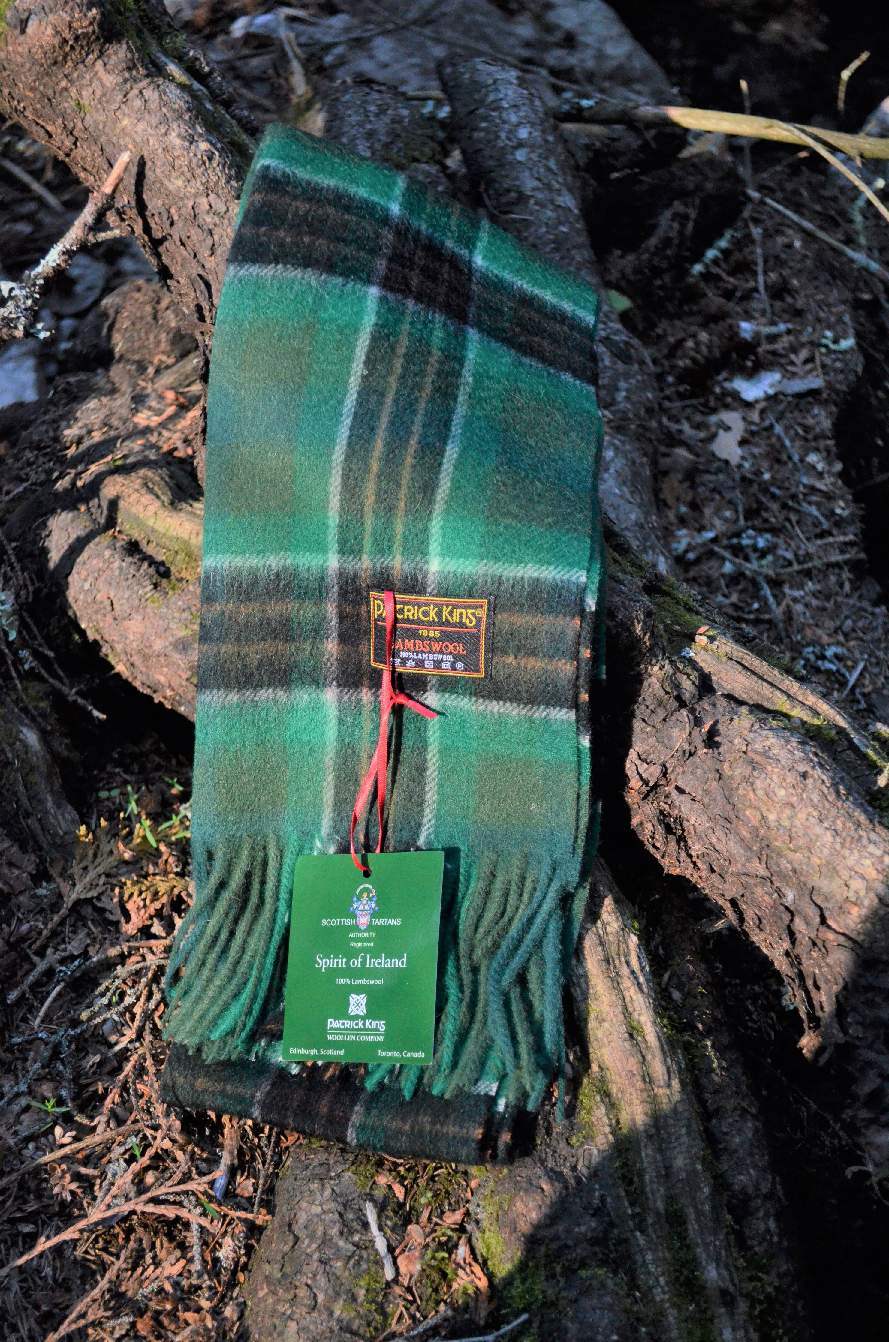 A Spirit of Ireland Lambswool Scarf by Patrick King, featuring a green tartan pattern and tag, is displayed on a rough tree trunk outdoors.
