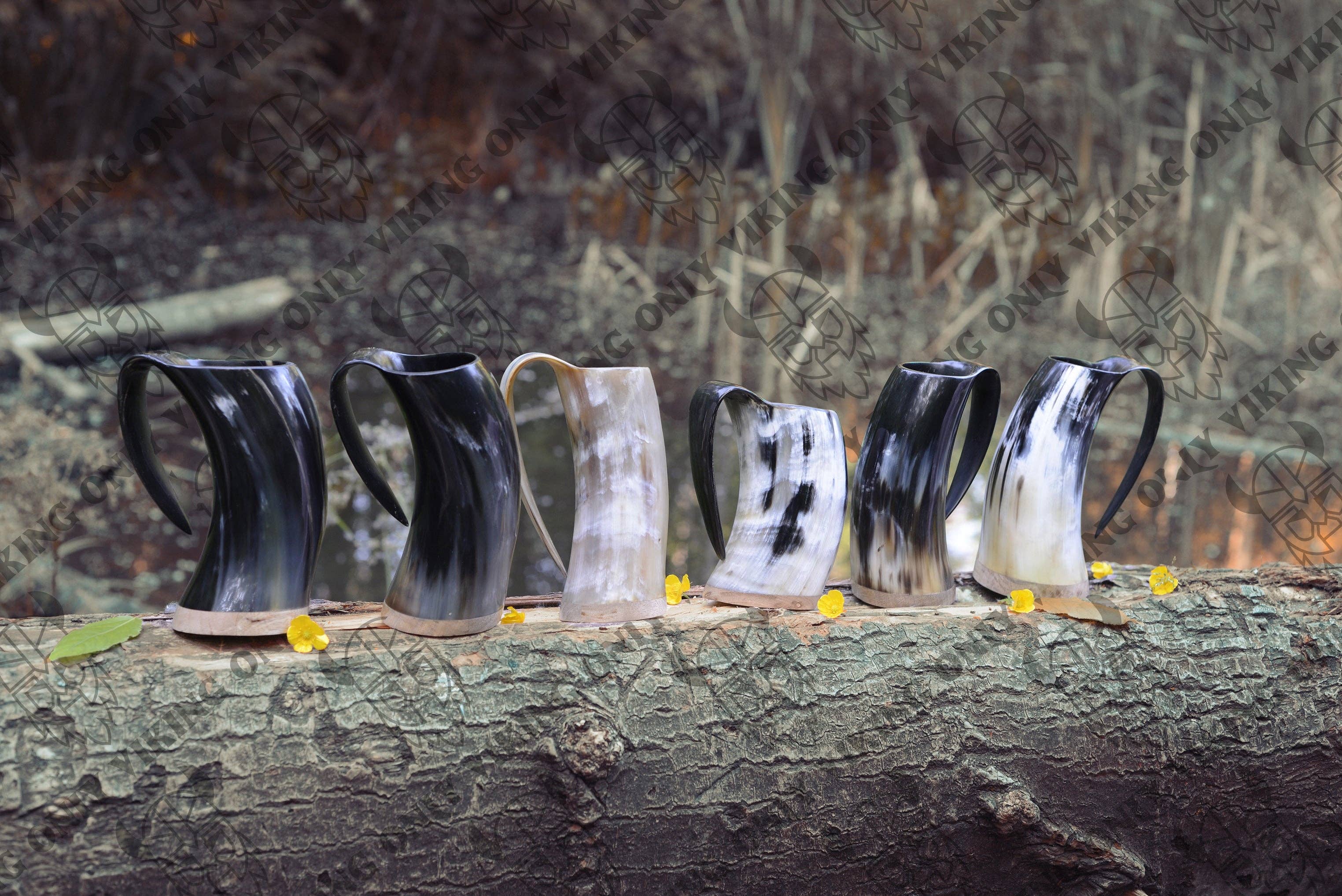 A row of Horn Mugs: Viking Ox Horn Coffee Mug 10oz, each showcasing unique patterns and colors, rests on a fallen tree trunk amidst small yellow flowers.