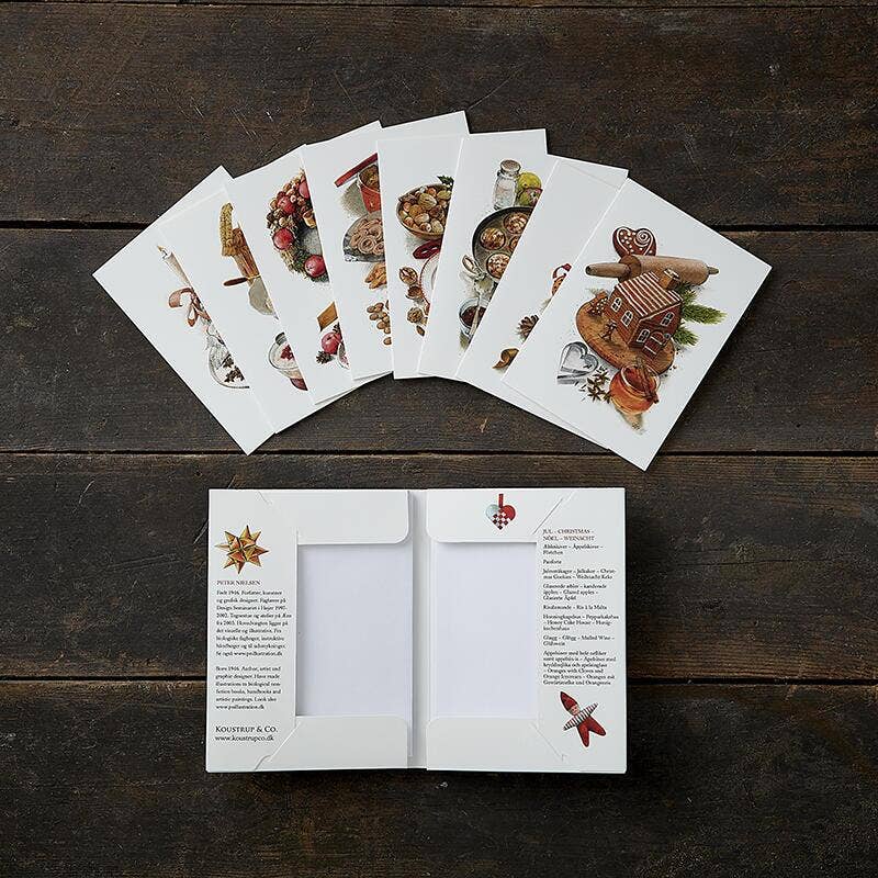 The "Card Pack: Jul A Taste of Christmas 8 Cards," featuring environmentally certified holiday cards with festive food and Christmas motifs, is displayed on a wooden table. One double card is opened up, showcasing the printed text and design elements.