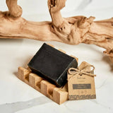 Black soap bar on a bamboo holder with a brown label tag tied in twine; large textured driftwood piece in the background.