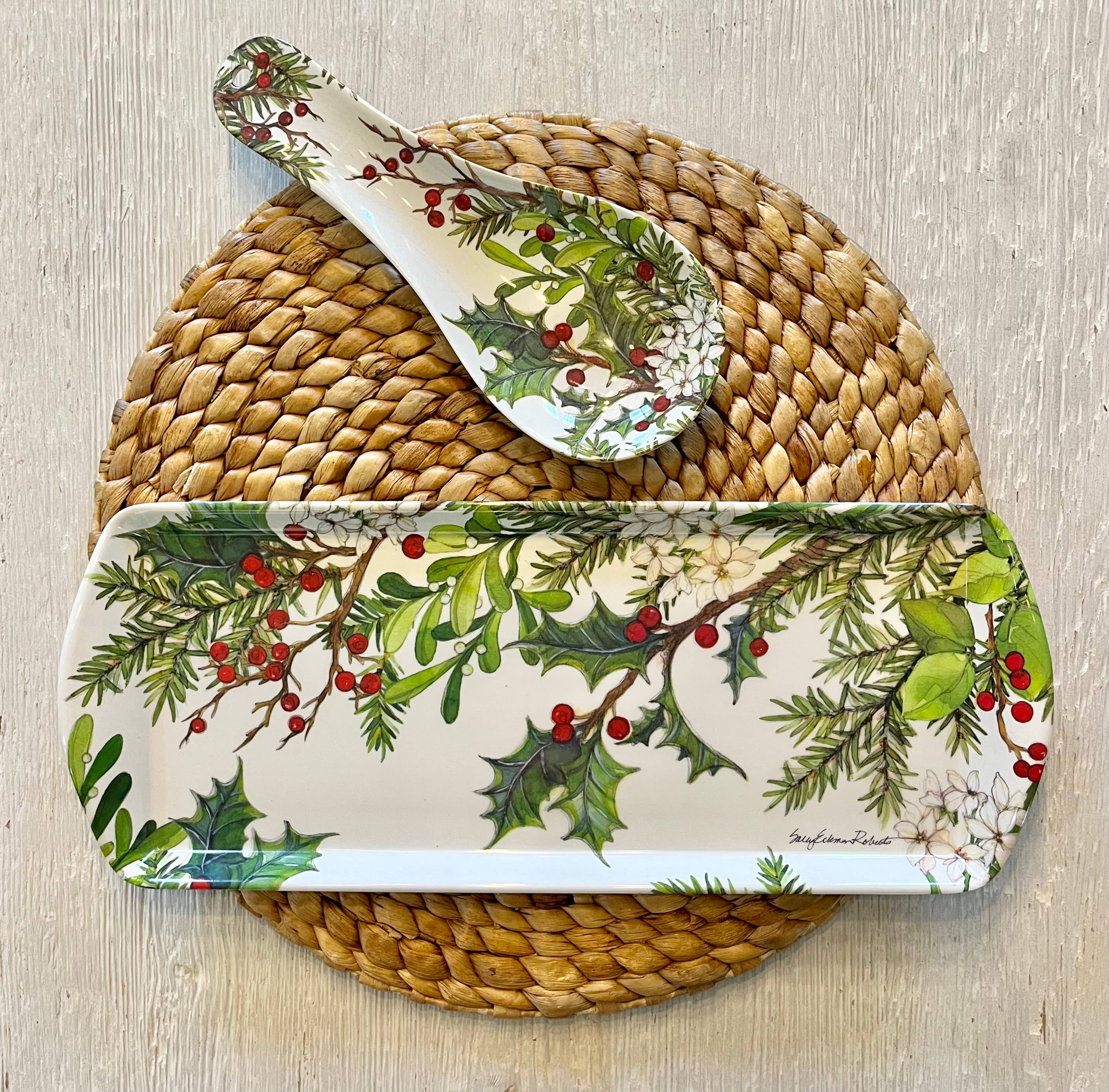 The Holly & Berries Holiday Serving Appetizer Tray, featuring festive foliage designs, sits atop a woven placemat on a light wood surface with a matching spoon rest.