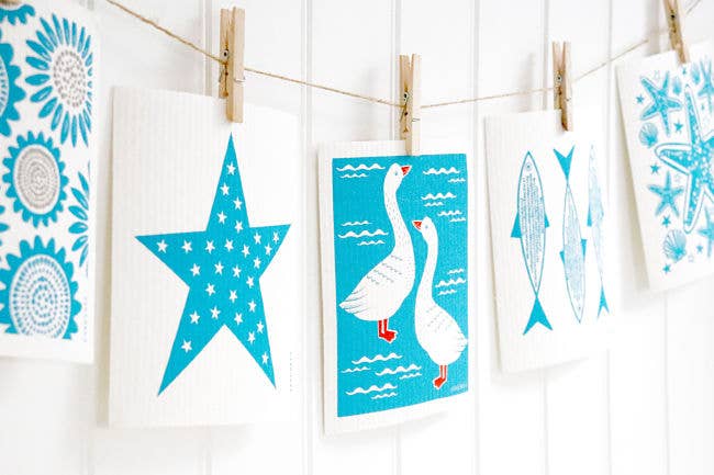 The biodegradable Swedish Dish Cloth in turquoise, featuring geese designs, hangs with wooden clothespins against a white backdrop, embodying charming Scandinavian style.