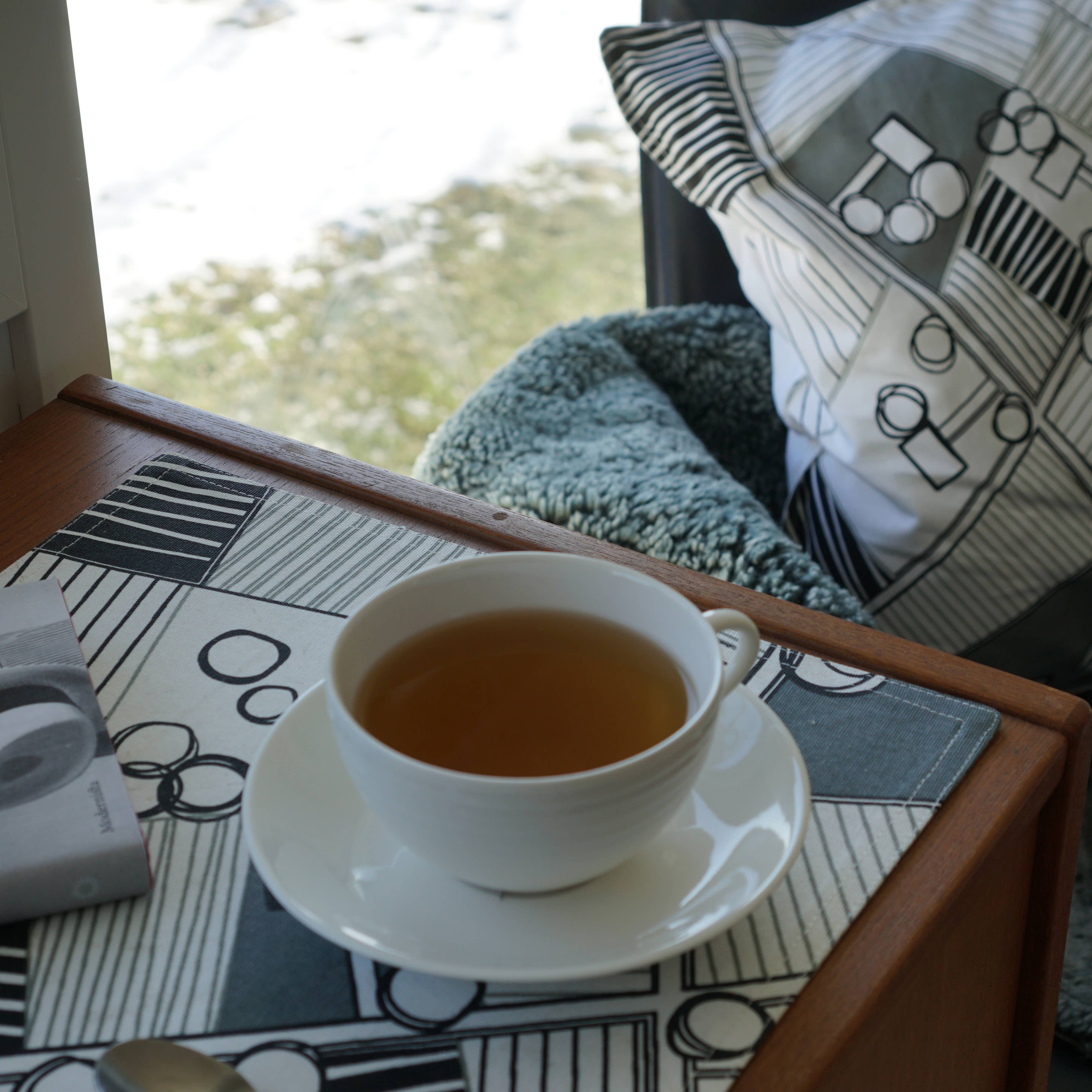 A cup of tea rests on a Born in Sweden double-sided placemat, measuring 47cm x 34cm, set beside a book. Nearby, a pillow and blanket adorn a chair by the window framing a snowy landscape.