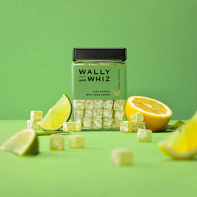 A jar of Wally & Whiz Lime w/Sour Lemon (240g) sits among candy cubes, a lemon half, and lime wedges on a green background—highlighting Danish candy with lime pieces coated in sour lemon flavor.