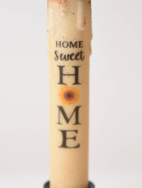 The Candle: Home Sweet Home Sunflower LED Timer Taper Candle features a cream color, sunflower image, and "Home Sweet Home" text, adding charm and convenience to your space with its top-dusted LED timer design.