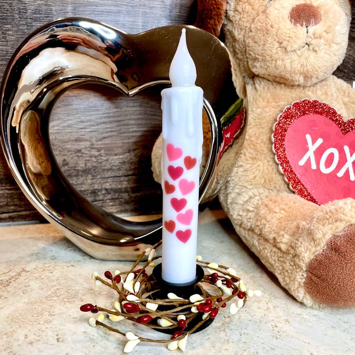 A decorative candle featuring red heart designs is elegantly displayed in the Square Wrought Iron Taper Holder, encircled by a ring of tiny berries. In the background, a plush bear with an "XOXO" heart and a metal heart sculpture enhance the charm of this delightful set-up.