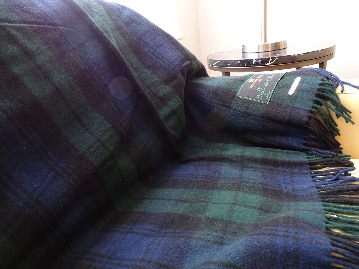 A Black Watch Highland "Tartan" Plaid Wool Blanket 69"x62" in dark green and navy is draped over a couch, with a small round side table and lamp visible in the background.