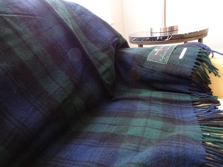 A Black Watch Highland "Tartan" Plaid Wool Blanket 69"x62" in dark green and navy is draped over a couch, with a small round side table and lamp visible in the background.