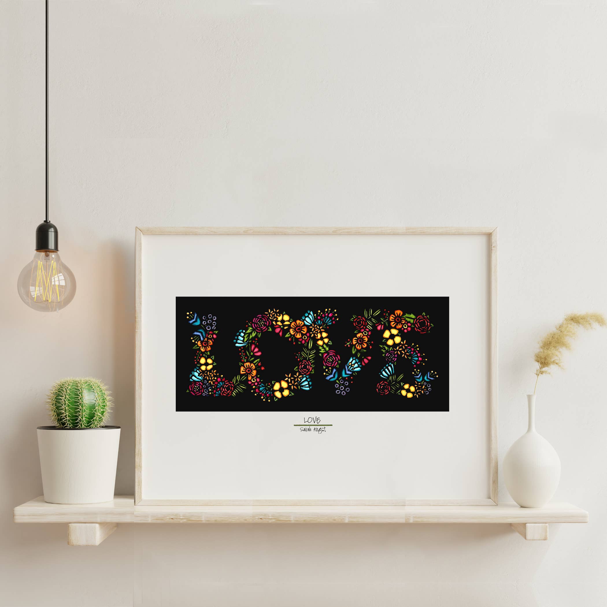 The 8x10 giclee print Artwork: LOVE Print by Sarah Angst features the word LOVE formed from colorful flowers and decorates a shelf with a cactus, pampas grass, and a hanging bulb to add charm to any space.