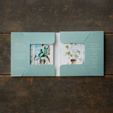 An open green cardfolder, made of FSC paper, lies on a wooden surface. It displays plant and bird illustrations near window motifs and is from the Card Pack: Room with a View—Square Cardfolder w/8 Cards & Envelopes collection.