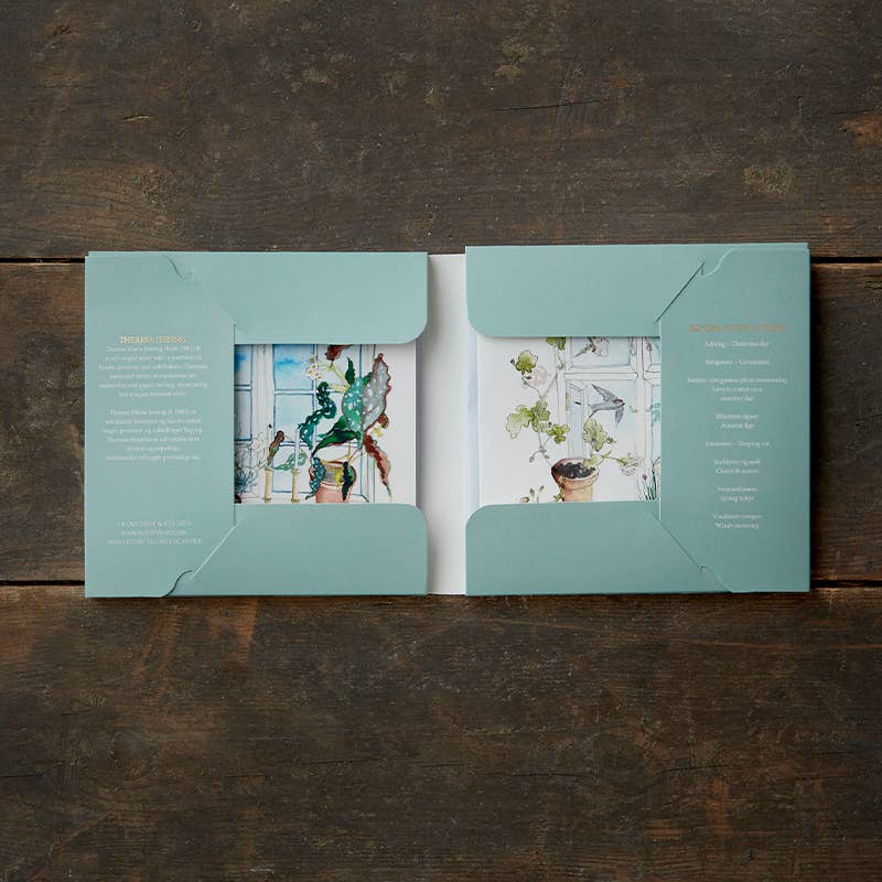 A square card pack, titled "Room with a view," rests on a wooden surface. The left page features artistic illustrations of leaves and birds, while the right page contains text. This booklet-style card folder is encased in a pale green cover and includes eight cards with envelopes.