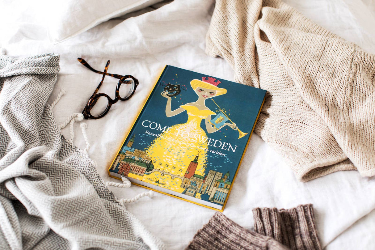 The Come to Sweden coffee table book, available in Swedish hardcover, rests on a bed with cozy blankets and glasses, ideal for vintage travel poster enthusiasts.