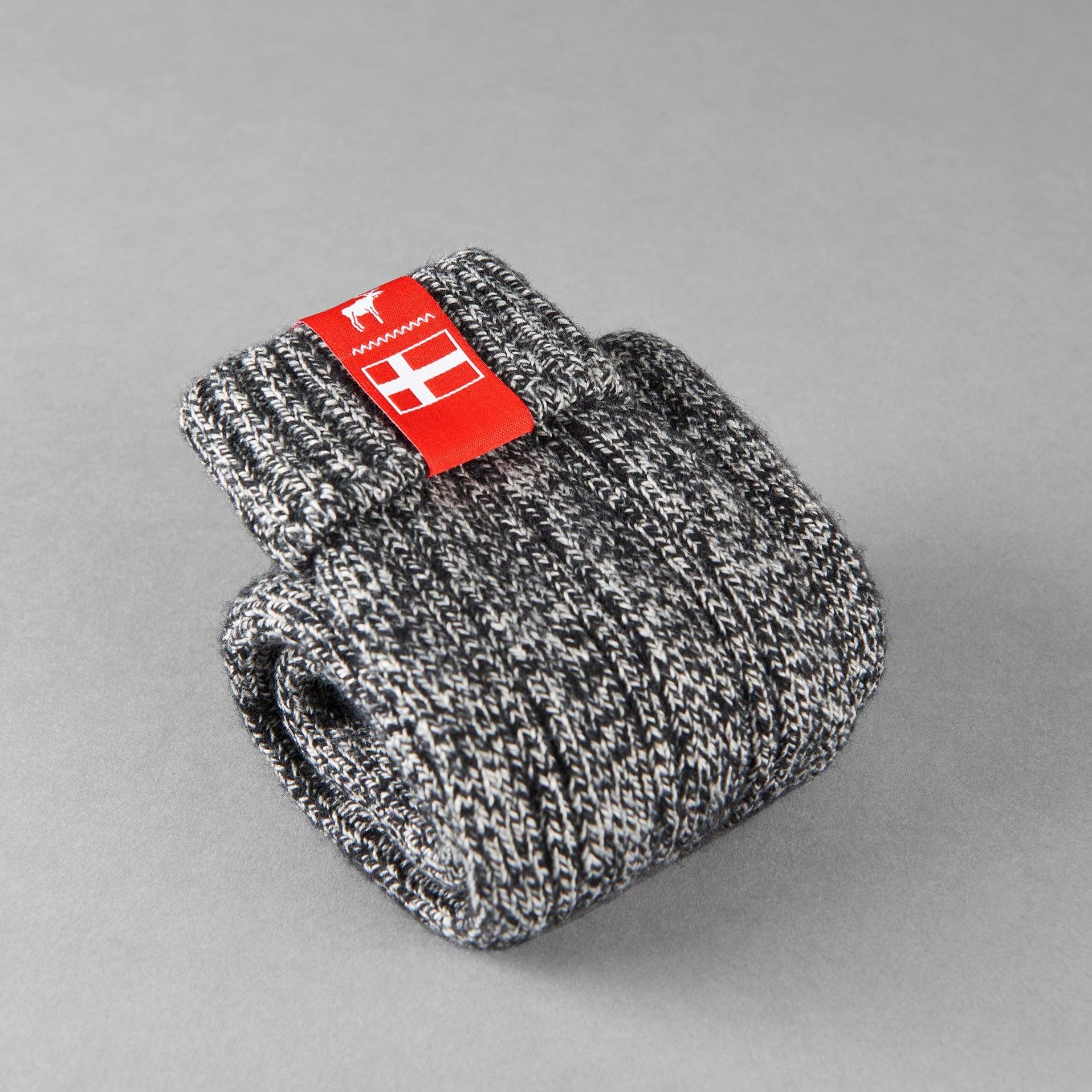 The Danish Hygge Socks - Nordic Noir, with their gray knitted wool design adorned by a red label and neatly folded against a gray background, capture the true spirit of comfort. Ideal for cozy winter evenings, these socks deliver warmth and coziness in every stitch.