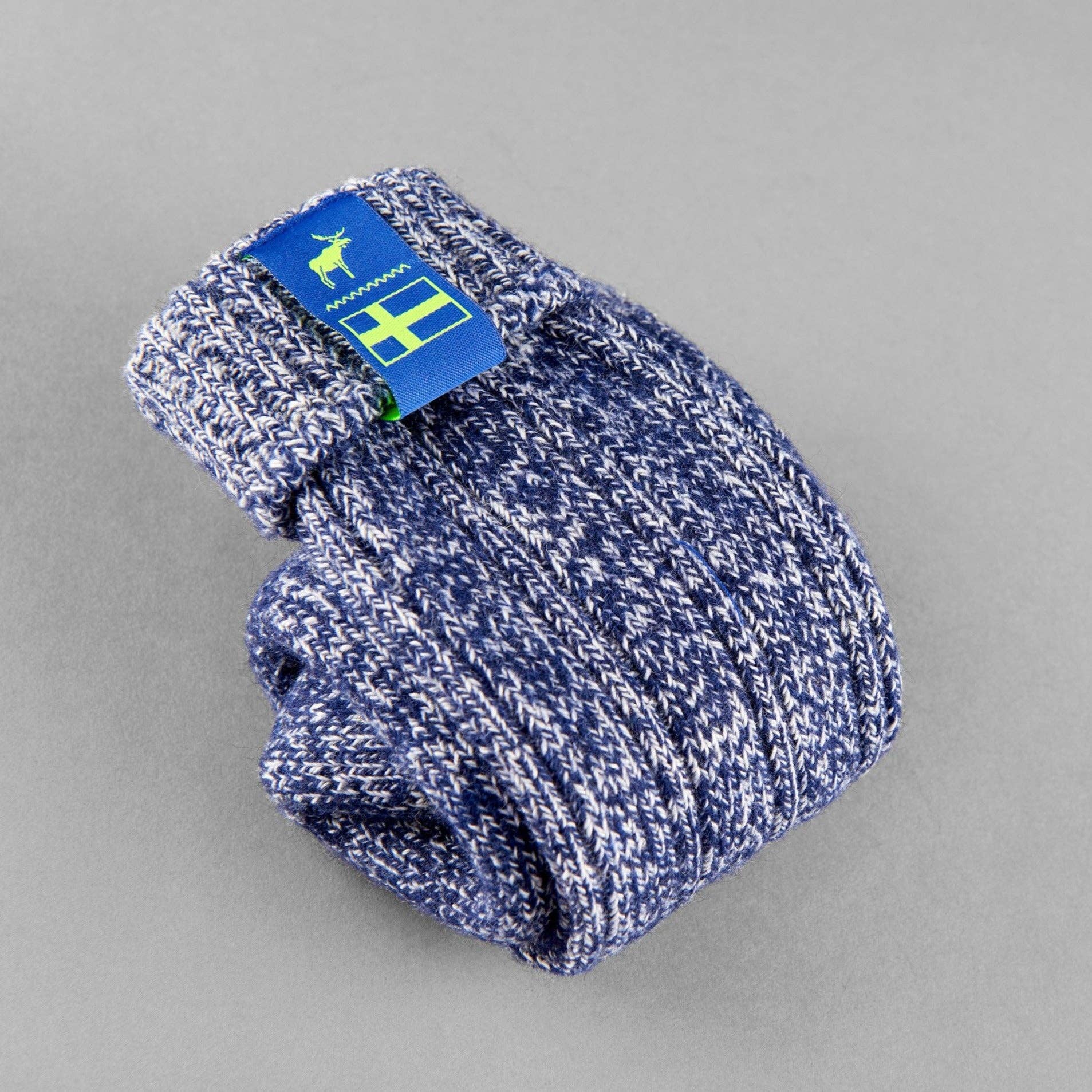 A pair of folded Classic Nordic Socks by The Nordic Sock Company in Swedish Midnight Blue, with a blue tag featuring a deer and green-yellow accents, displayed on a plain gray background.